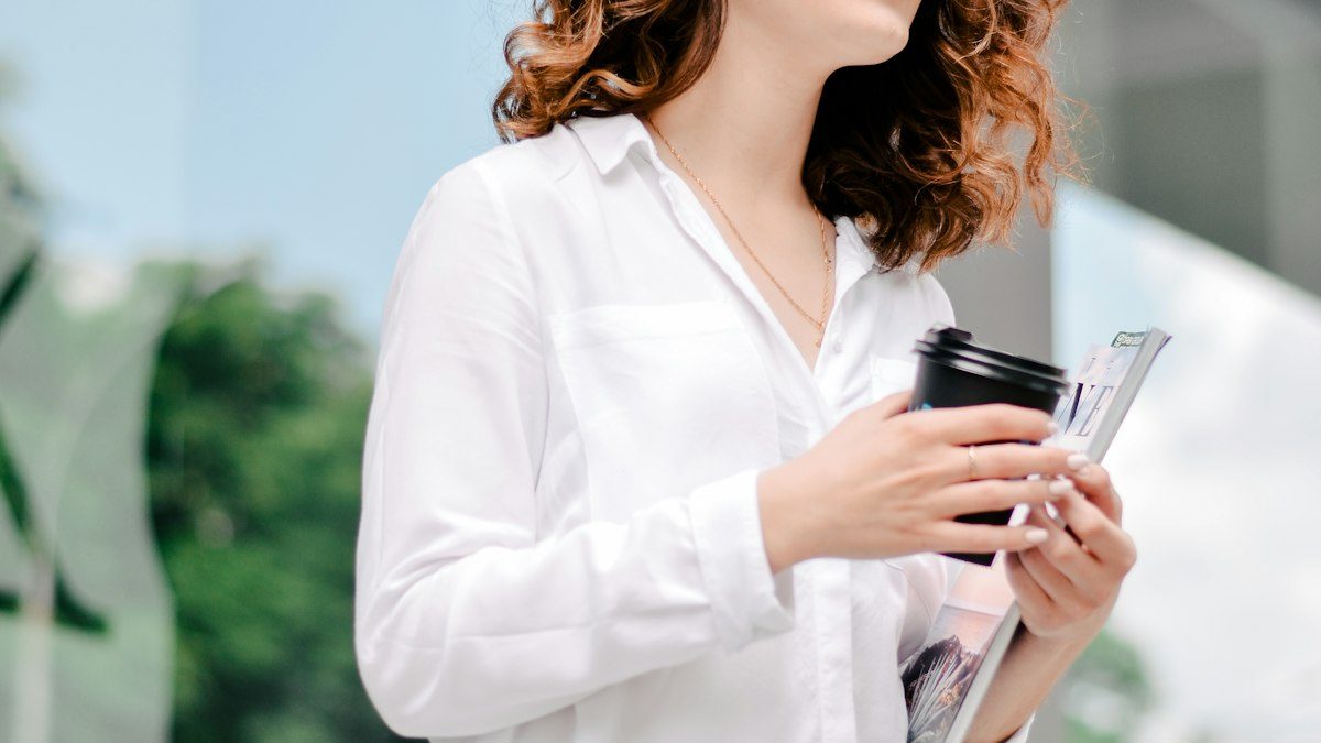 Mujer con camisa blanca sosteniendo un café y papeles en la calle